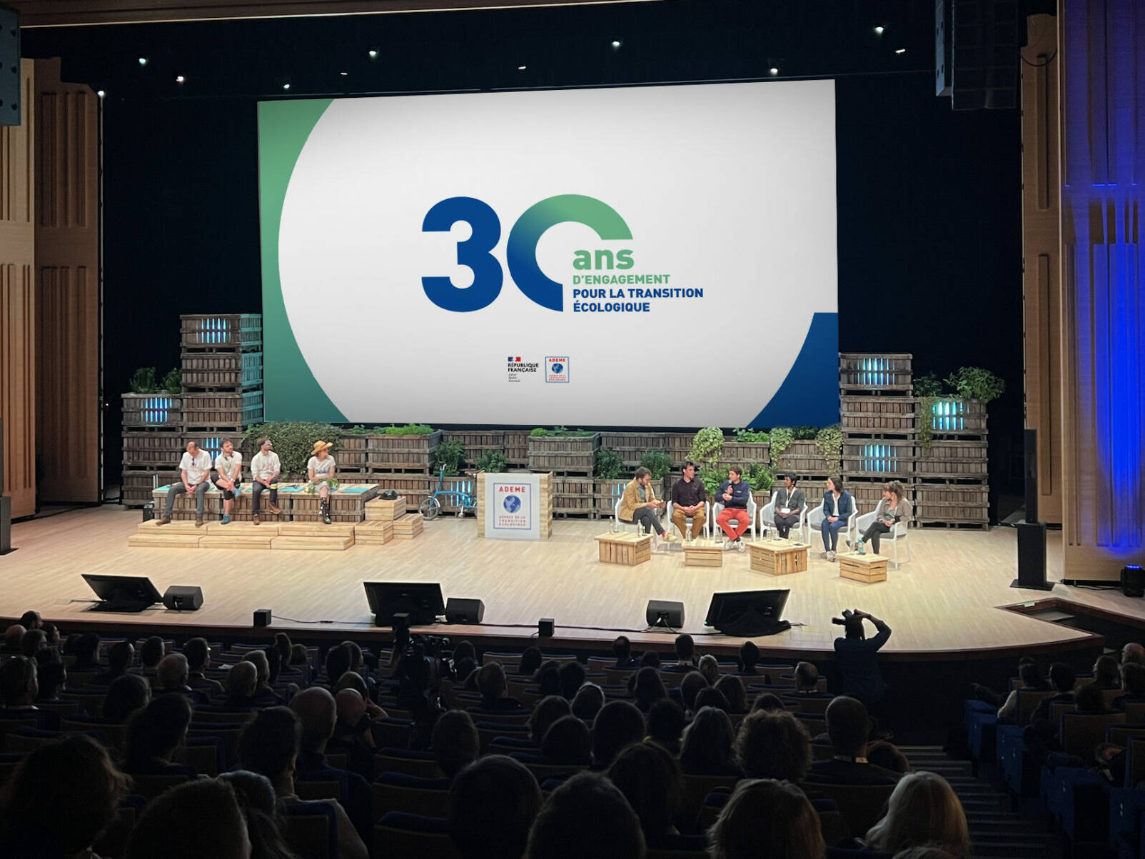 Conférence anniversaire de l’ADEME pour 30 ans d’engagement pour la transition écologique, avec intervenants sur scène, public en auditorium et décor événementiel en palettes de bois végétalisées.