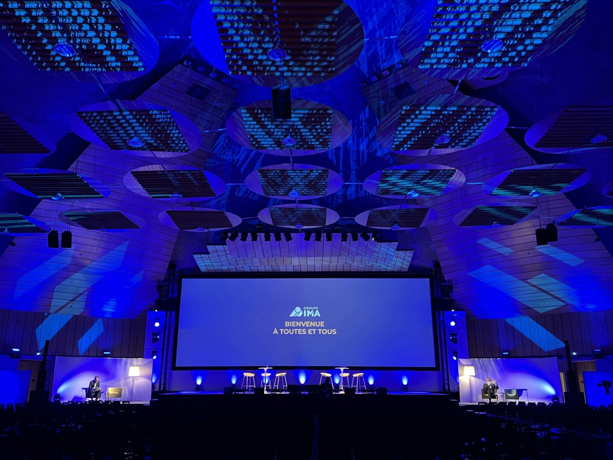 Salle de conférence JDM IMA avec écran géant, ambiance lumineuse bleue, scène aménagée pour une table ronde et scénographie immersive en auditorium.