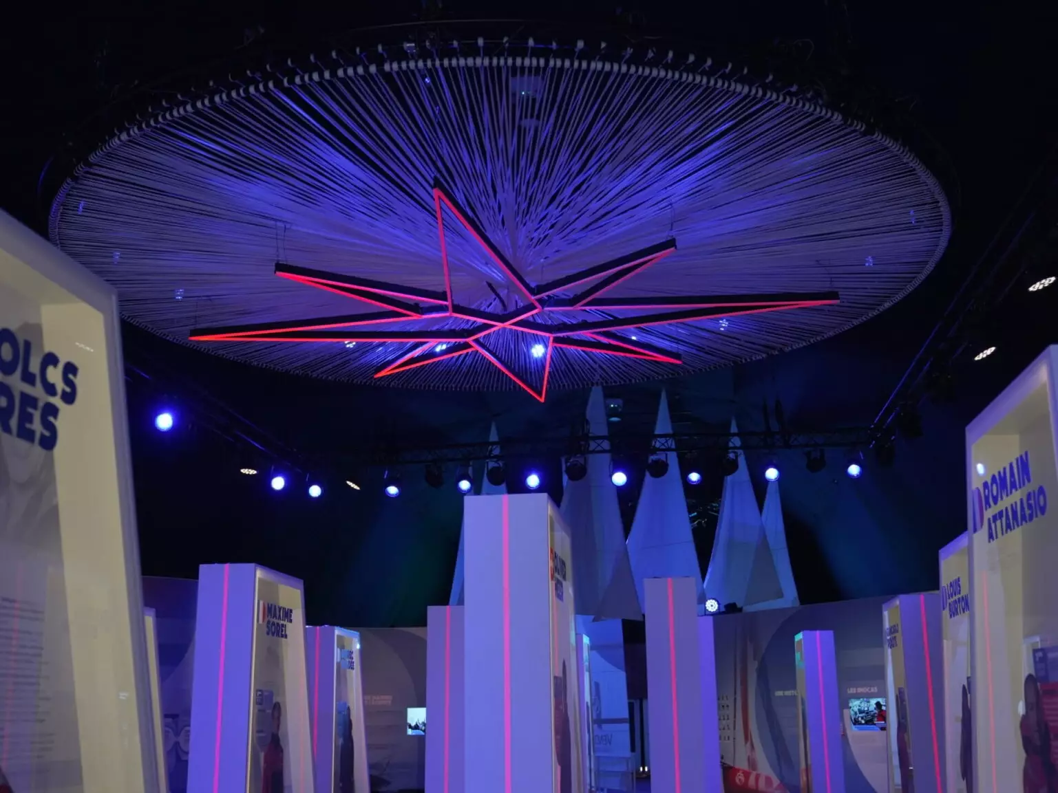 Scénographie immersive du Vendée Globe avec installation lumineuse suspendue, stands d’exposition et ambiance bleue événementielle.