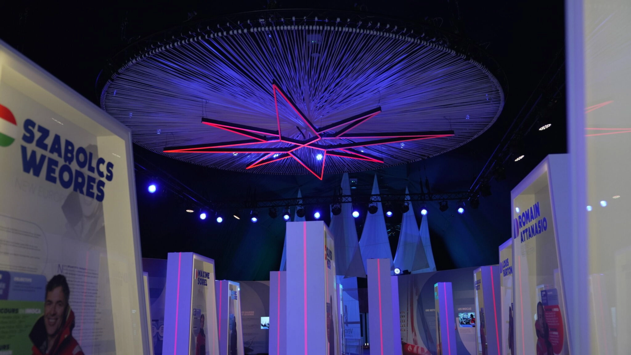 Scénographie immersive du Vendée Globe avec installation lumineuse suspendue, stands d’exposition et ambiance bleue événementielle.