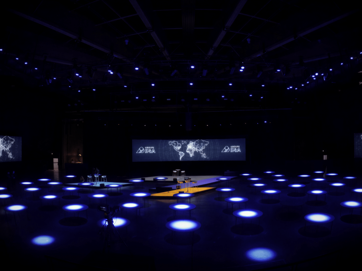 Salle événementielle plongée dans une ambiance bleue pour la JDM d'IMA, avec tables lumineuses, écrans panoramiques, projections graphiques et mise en lumière immersive.