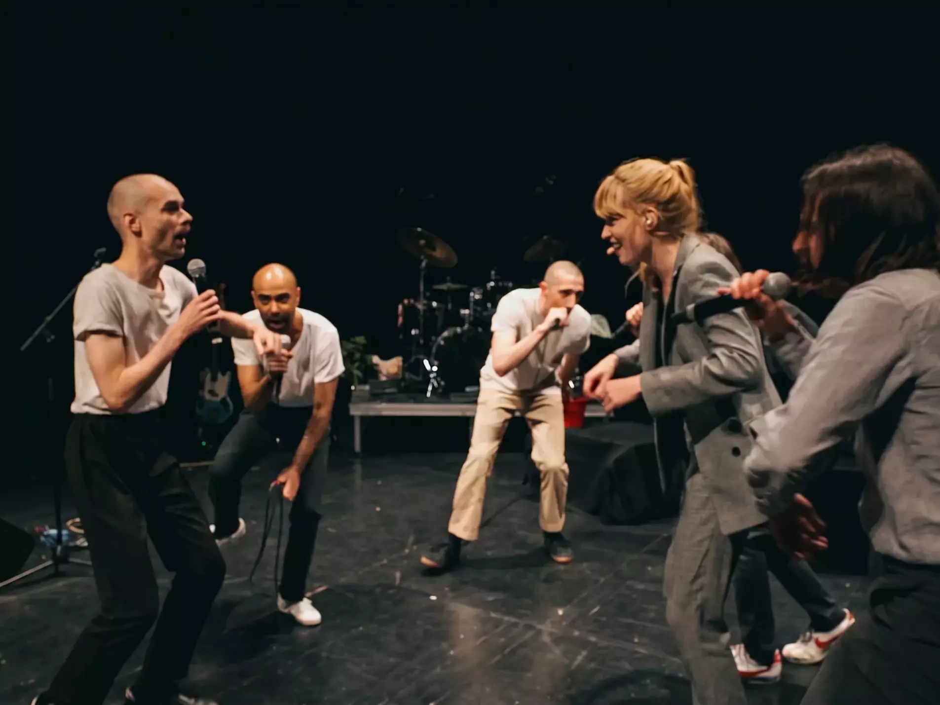 Groupe d’artistes en pleine performance live sur scène, avec micros, chant, jeu théâtral et instruments en arrière-plan dans un spectacle musical contemporain.